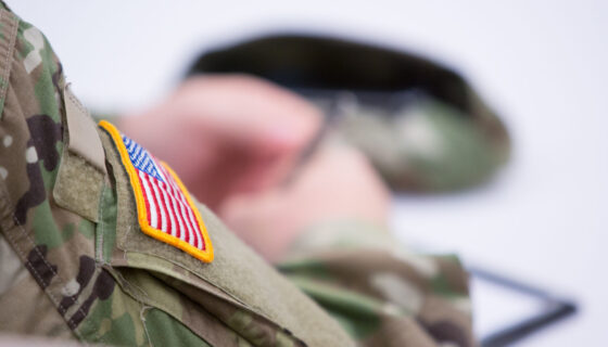 A close-up image of a person in military camouflage uniform with an American flag patch on the sleeve. The person's hands are blurred and holding an object, possibly a hat or cap, which is also partially visible in the background. The focus is on the uniform and the flag patch.