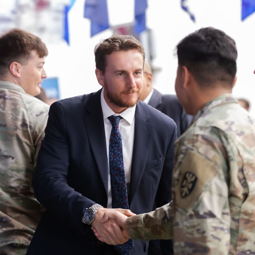 A man in a suit and tie shakes hands with a man in uniform with other uniformed service members in the background.